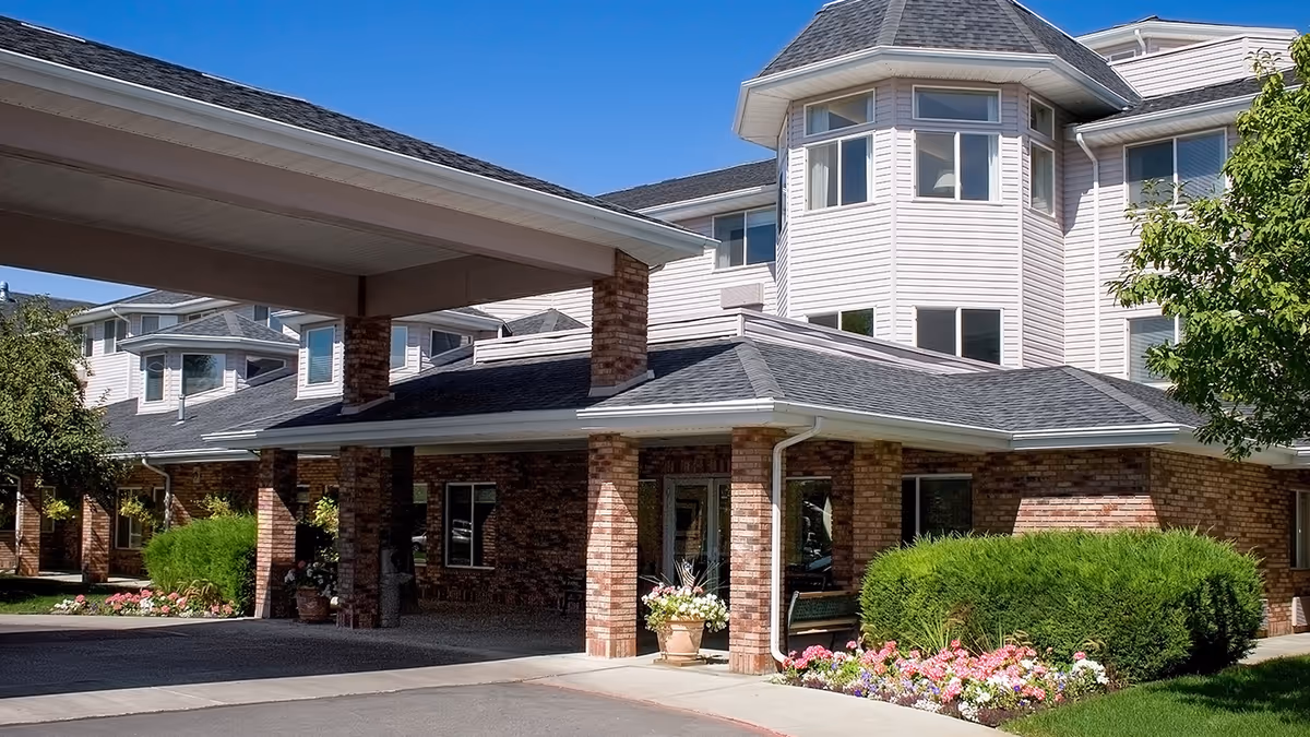 Exterior view of Holiday Aspen View facility showing a covered entrance with brick pillars, manicured bushes, colorful flower beds, and a multi-story building with white siding and multiple windows under a clear blue sky.