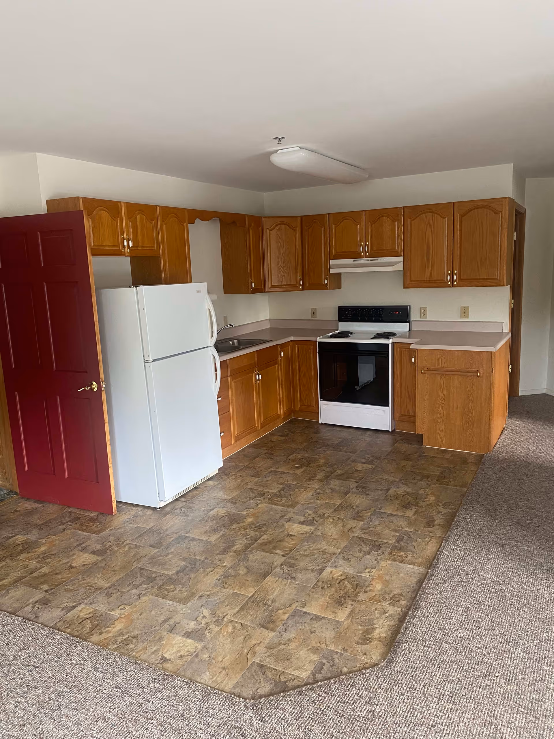 A kitchen area with wooden cabinets, a white refrigerator, a white stove with an overhead vent, and a stainless steel sink. The floor in the kitchen area is covered with patterned vinyl tiles, while the adjacent area has carpet flooring. A partially open red door is visible on the left side.