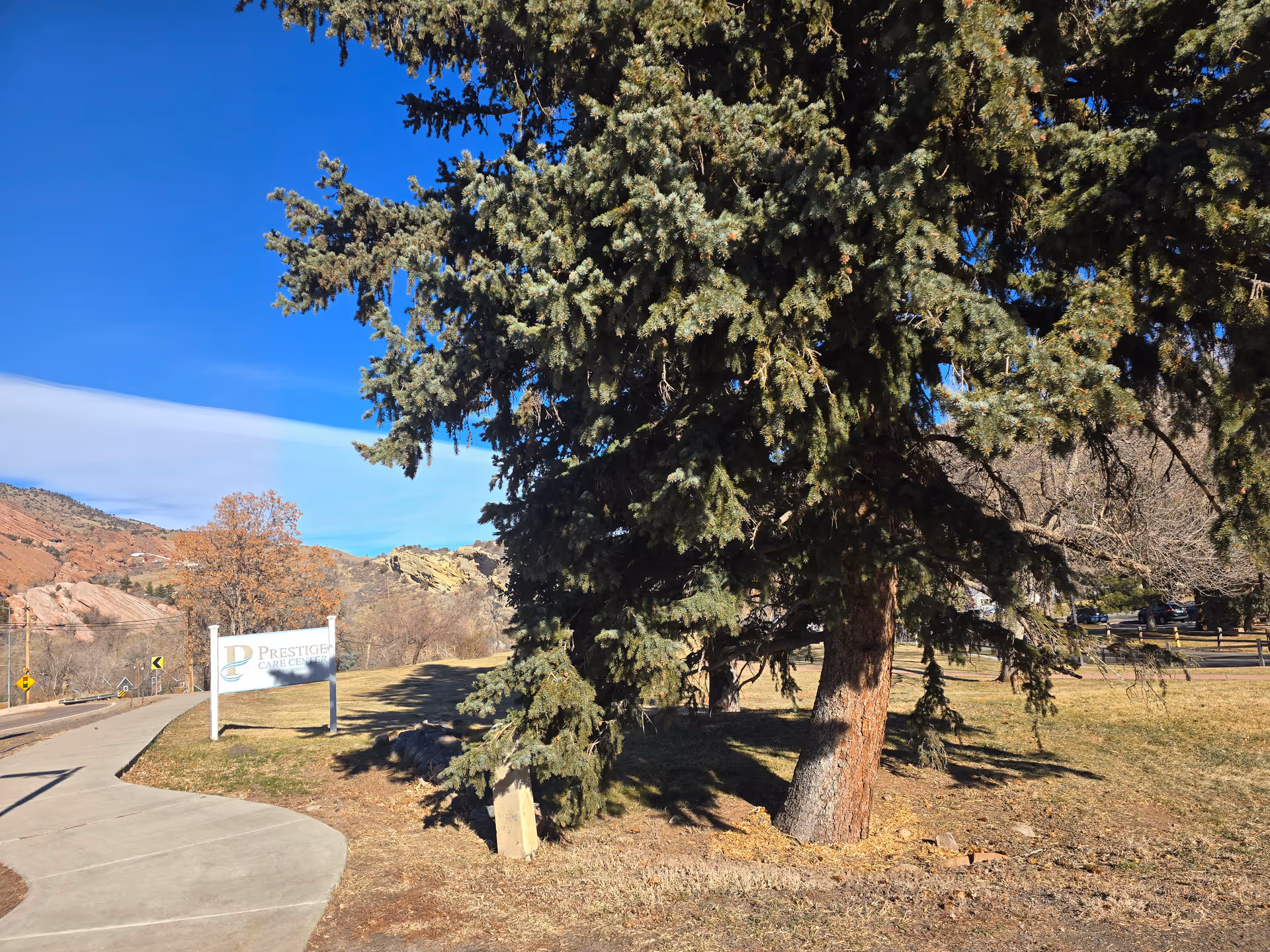 Outdoor view of a landscaped area with a large evergreen tree in the foreground and a sidewalk curving to the left. A sign reading 'Prestige Care Center' is visible on the grass near the sidewalk. Hills and a clear blue sky are in the background.