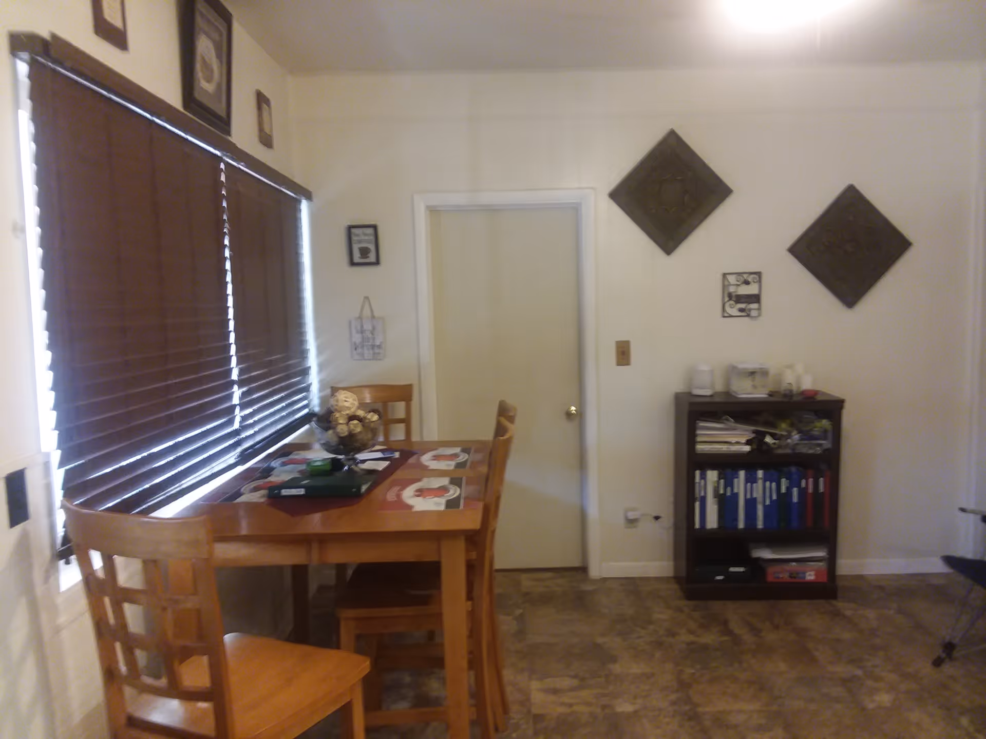 Interior view of a dining area with a wooden table and four chairs. The table has placemats and a decorative centerpiece. There are closed brown blinds covering the windows on the left wall. On the right wall, there is a small bookshelf with binders and some items on top, along with decorative wall hangings above it. A closed door is centered on the back wall.