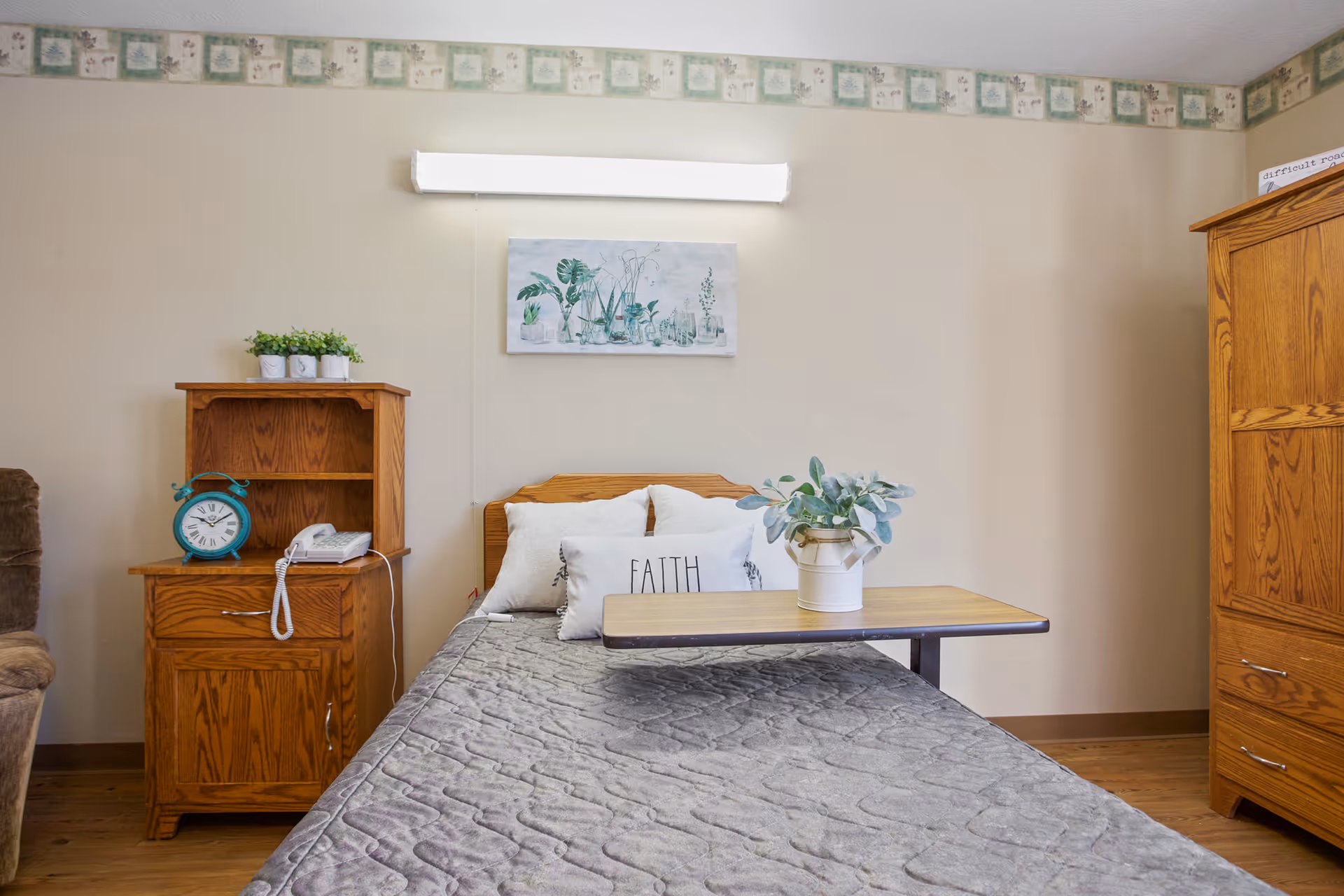 A single bed with a gray quilted blanket and white pillows, one pillow has the word 'FAITH' printed on it. A small wooden overbed table with a white vase holding green foliage is placed on the bed. To the left of the bed is a wooden nightstand with a blue alarm clock, a white telephone, and three small potted plants on top. On the right side of the image is a wooden wardrobe. The walls are beige with a floral border near the ceiling, and a botanical-themed painting hangs above the bed.