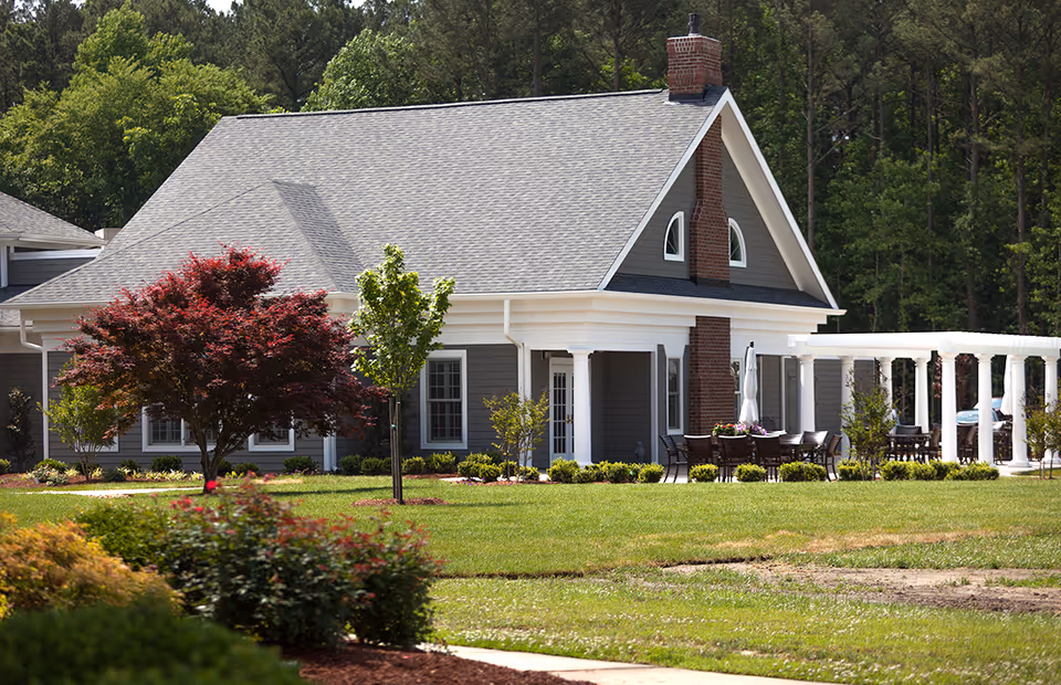 A single-story building with gray siding and a steep gray shingled roof, featuring a brick chimney and white trim. The building is surrounded by a well-maintained lawn with various trees and shrubs. There is an outdoor seating area with tables and chairs under a white pergola on the right side of the building. Tall trees form a wooded background.