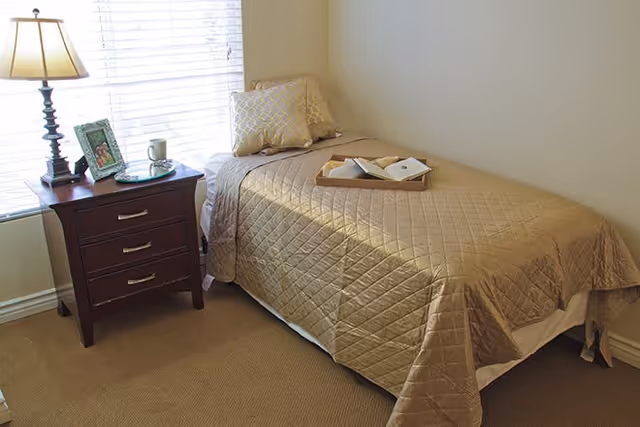 A small bedroom with a single bed covered in a beige quilted bedspread and two decorative pillows. A wooden nightstand with three drawers is beside the bed, holding a table lamp, a framed photo, and a cup. A window with white blinds is behind the nightstand.