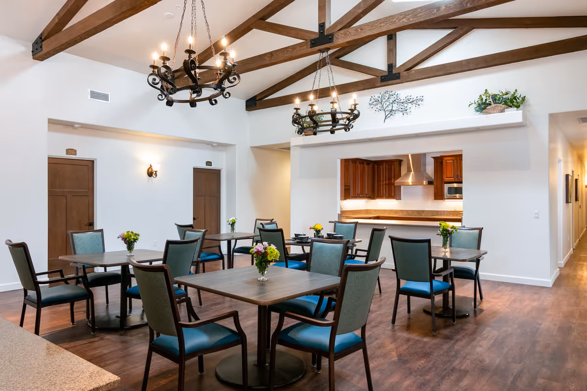 Open dining room with multiple tables and teal-upholstered chairs, exposed wood ceiling beams, chandeliers, and a serving kitchen in the background.