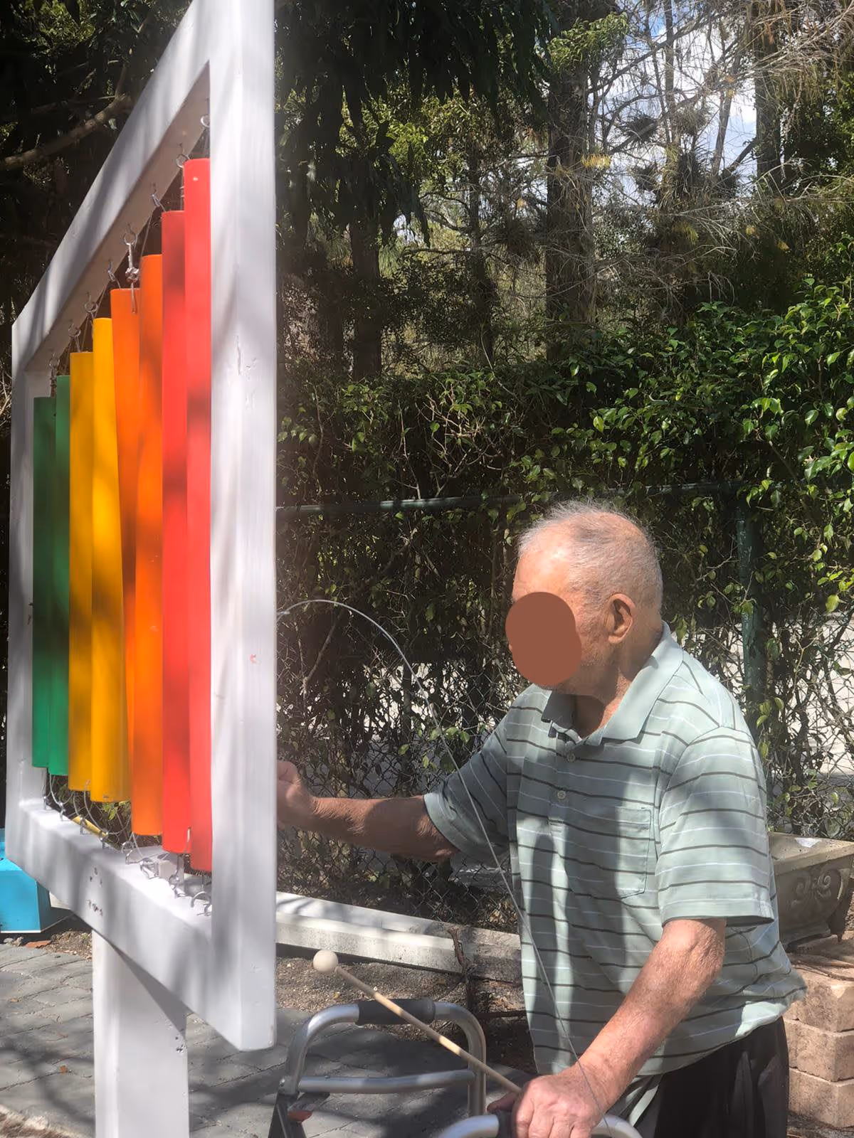 An elderly man using a walker plays a large outdoor xylophone with colorful bars in a garden area surrounded by trees and greenery.