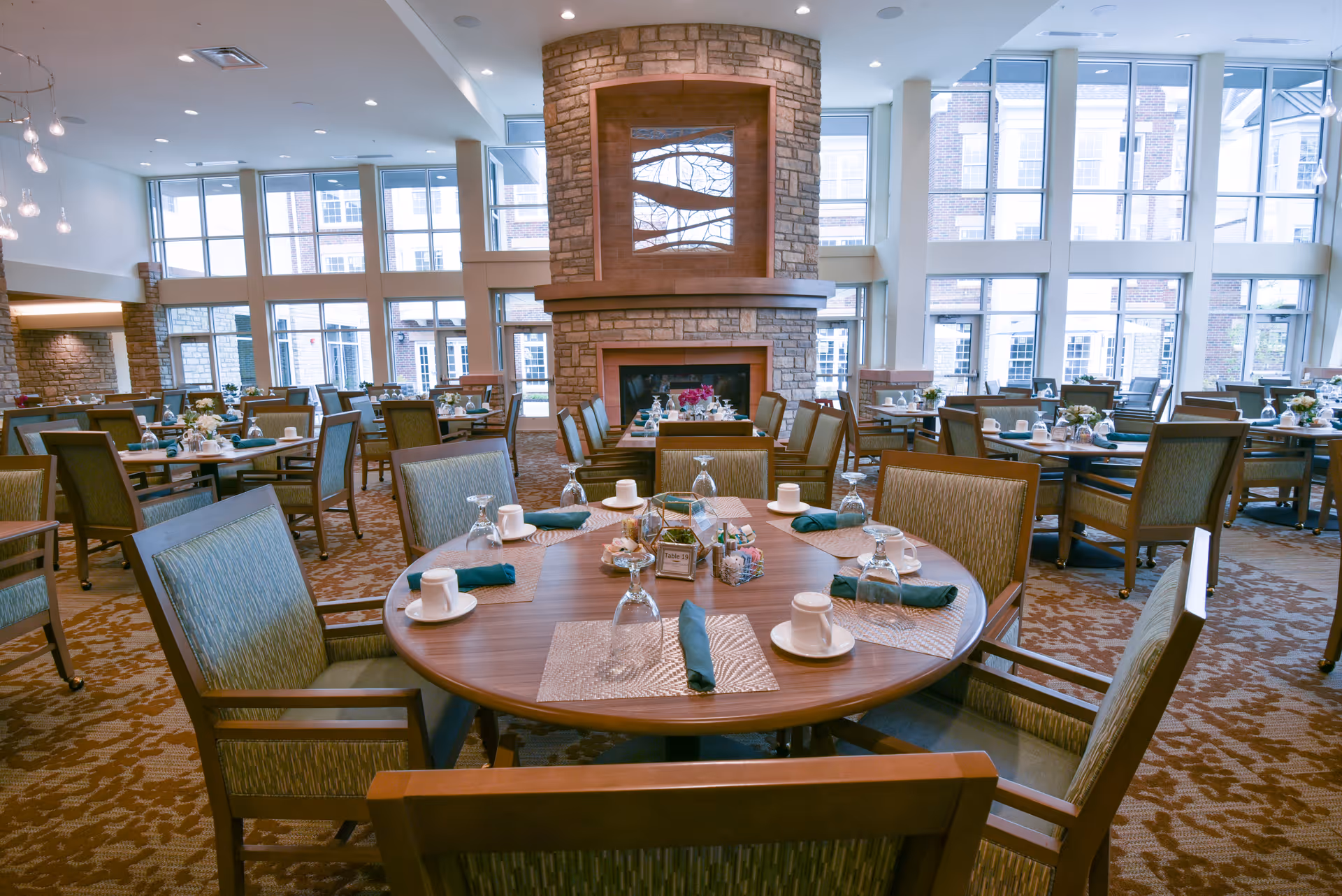 A spacious dining room with multiple round wooden tables set with placemats, cups, napkins, and upside-down glasses. The room features large windows allowing natural light to fill the space, and a stone fireplace with a decorative wooden frame is centrally located. Comfortable upholstered chairs surround each table, and the carpet has a patterned design.