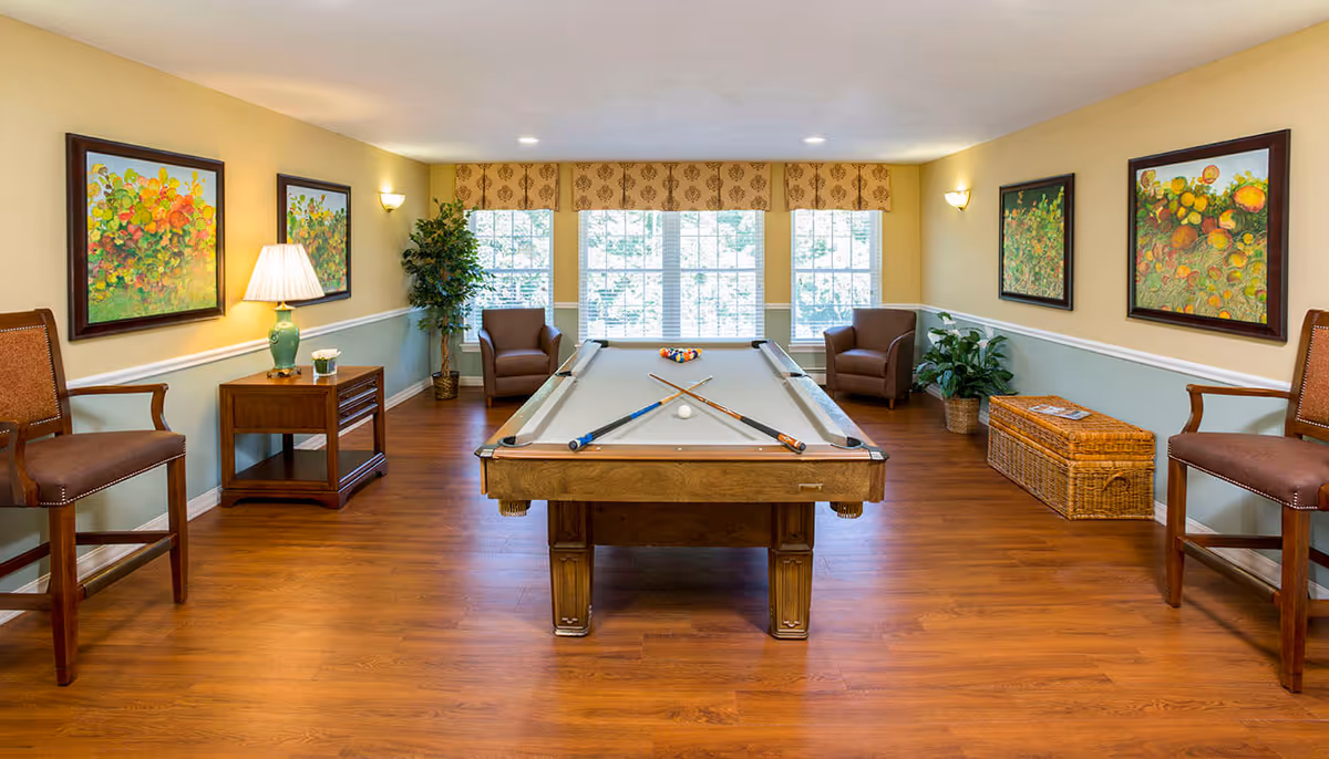A well-lit recreational room with a pool table centered in the middle. The pool table has two crossed cues and a white cue ball on its beige felt surface, with a rack of colorful billiard balls at the far end. The room features wooden flooring, two brown armchairs near large windows with white blinds and a patterned valance, and several framed colorful paintings on the light yellow walls. There are also two wooden chairs, a small wooden side table with a green lamp, and some potted plants and a wicker storage basket along the walls.