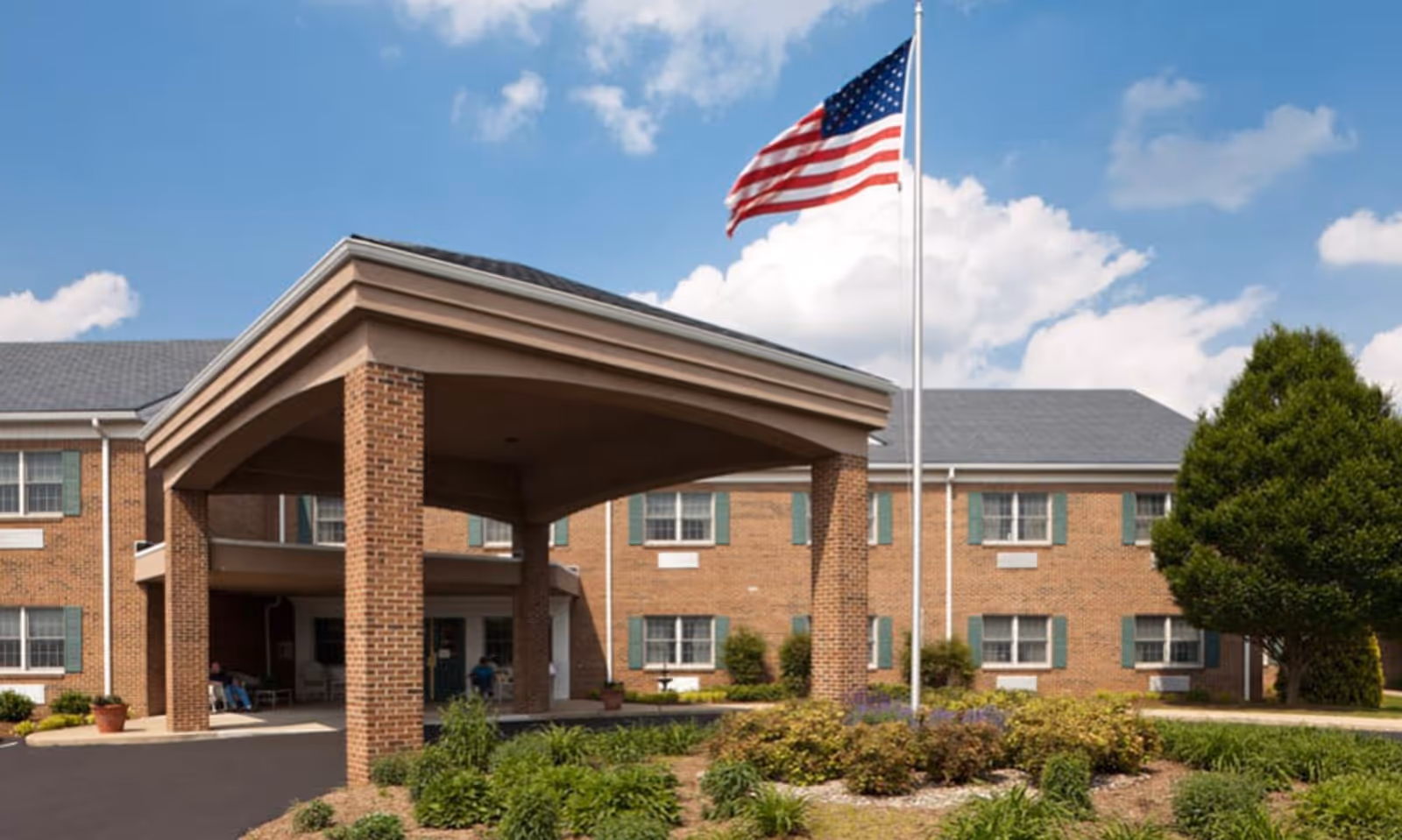 Exterior view of a two-story brick senior living facility with green shutters, a covered entrance supported by brick columns, an American flag on a flagpole, and landscaped greenery in front under a partly cloudy blue sky.