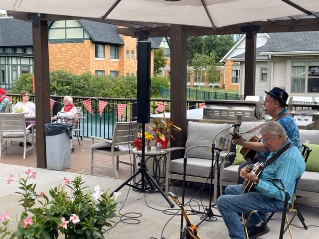 Two musicians playing guitars on an outdoor patio under a pergola while residents sit at tables near a pool with brick buildings in the background.