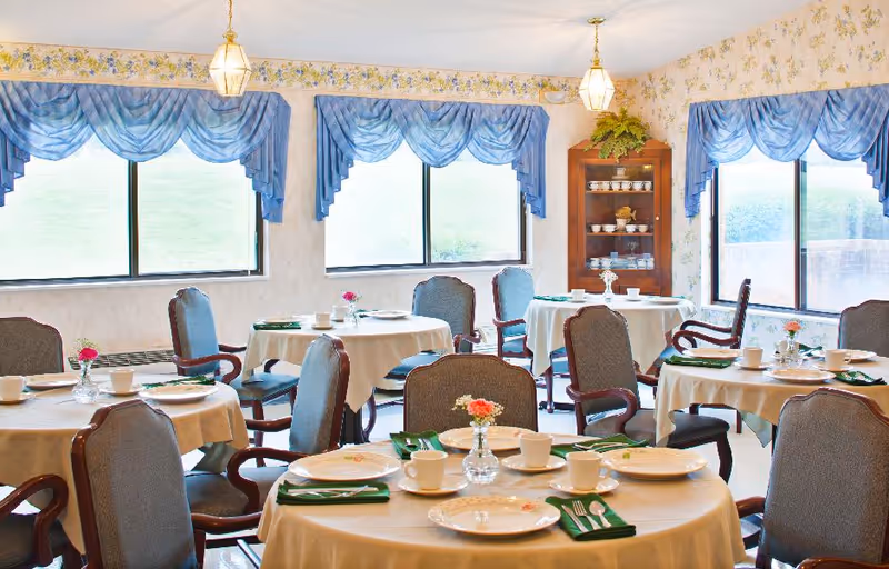Bright dining room with round tables set for meals, upholstered chairs, blue valance curtains, and a china cabinet.