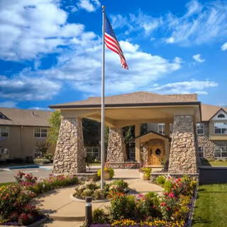 Entrance of a senior living facility with a covered driveway supported by stone pillars, an American flag on a flagpole in front, and landscaped flower beds along the walkway under a partly cloudy blue sky.