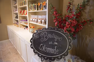 Interior view of a boutique area in a senior living facility featuring a chalkboard sign that reads 'The Boutique at the Colonnade', shelves stocked with various packaged goods, and a vase with tall pink flowers on a table.