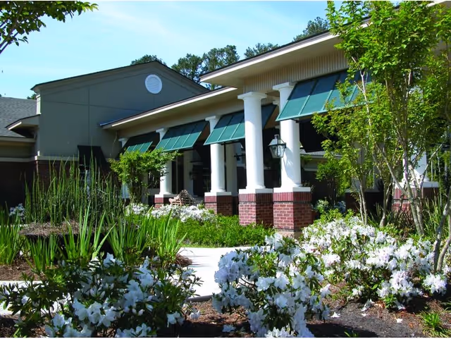 Front entrance of a nursing facility with white columns, green awnings, and landscaped flower beds.