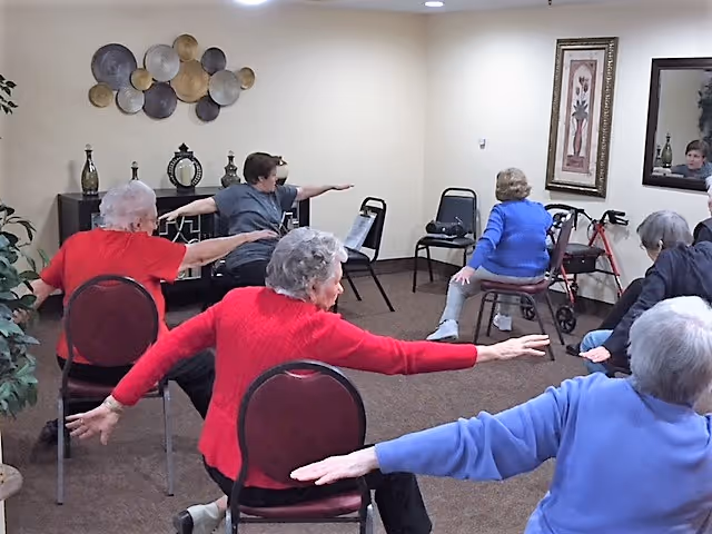 A group of elderly individuals seated on chairs in a room, participating in a seated exercise session with arms extended. The room has beige walls, decorative wall art, and a mirror. Some mobility aids like a walker are visible.