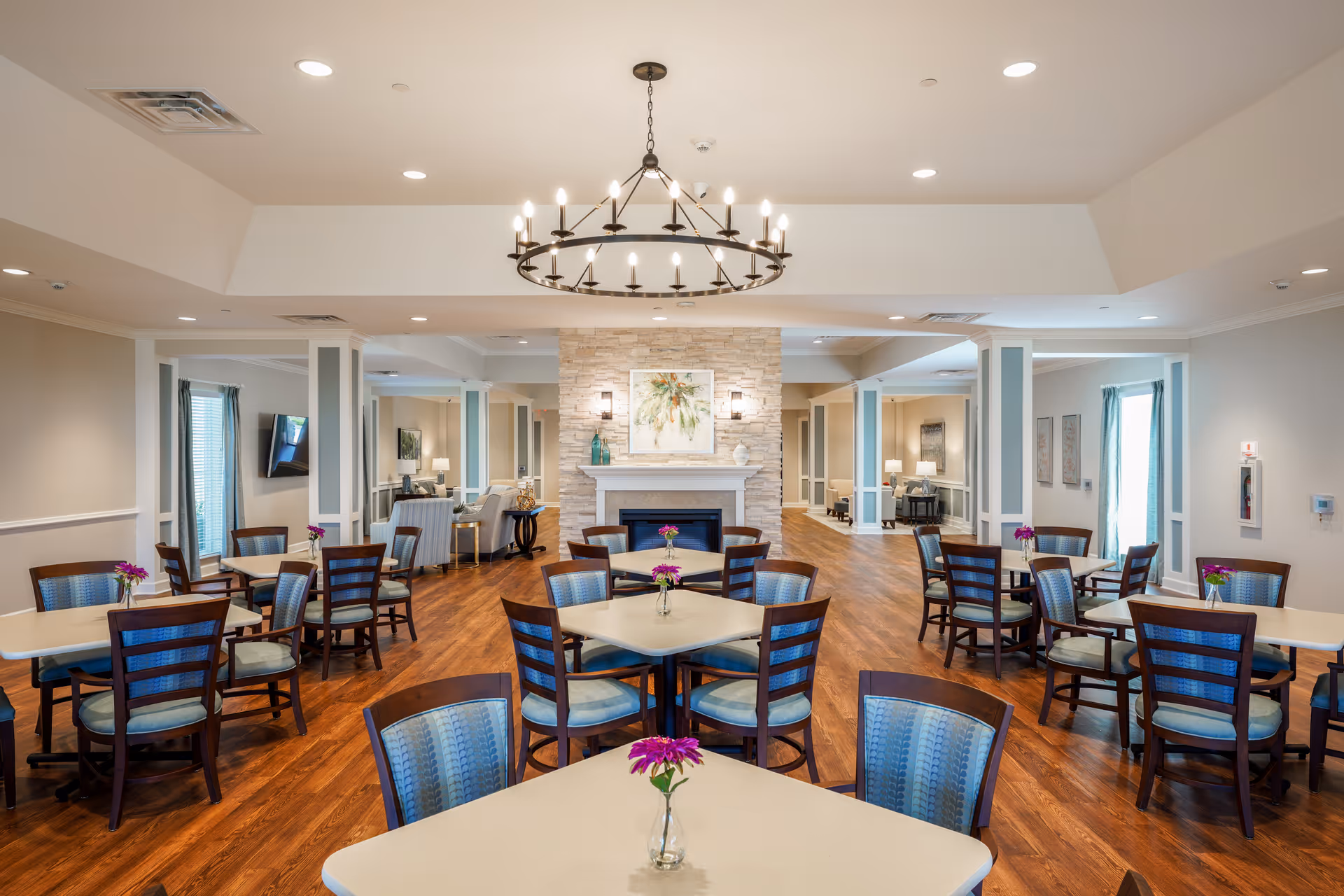 A spacious dining room in an assisted living facility with multiple square tables, each surrounded by wooden chairs with blue cushioned seats. Each table has a small vase with purple flowers. The room features wood flooring, a stone fireplace with a painting above it, and a large circular chandelier hanging from the ceiling. There are windows with blue curtains and additional seating areas visible in the background.