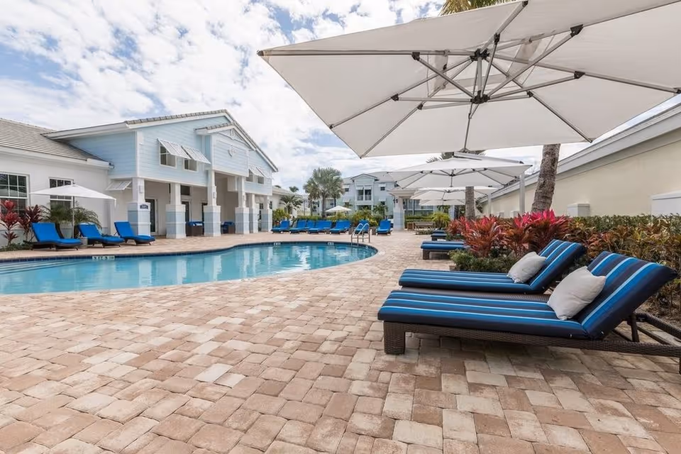 Outdoor pool area at a senior living facility with blue cushioned lounge chairs, large white umbrellas, and a clear blue swimming pool surrounded by a brick-paved deck. The background shows light blue and white buildings with palm trees and partly cloudy sky.