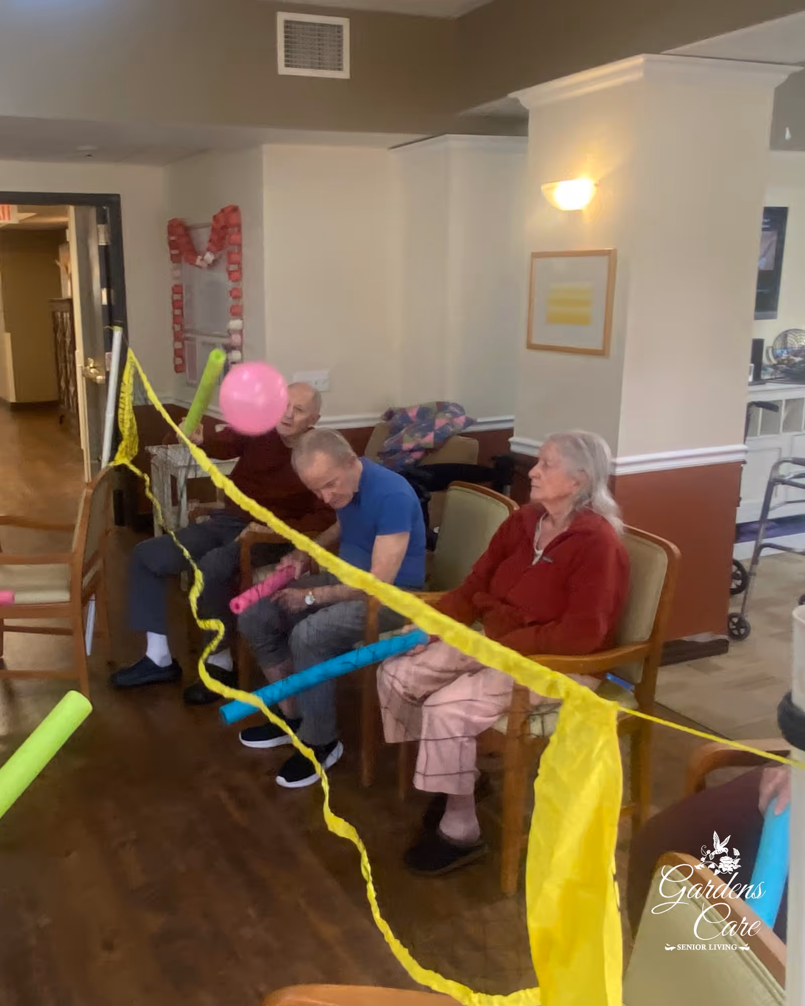 Three elderly individuals seated in chairs indoors playing a balloon volleyball game using pool noodles and a makeshift net made of yellow tape and netting. The room has light-colored walls and wooden flooring, with some furniture and a walker visible in the background.