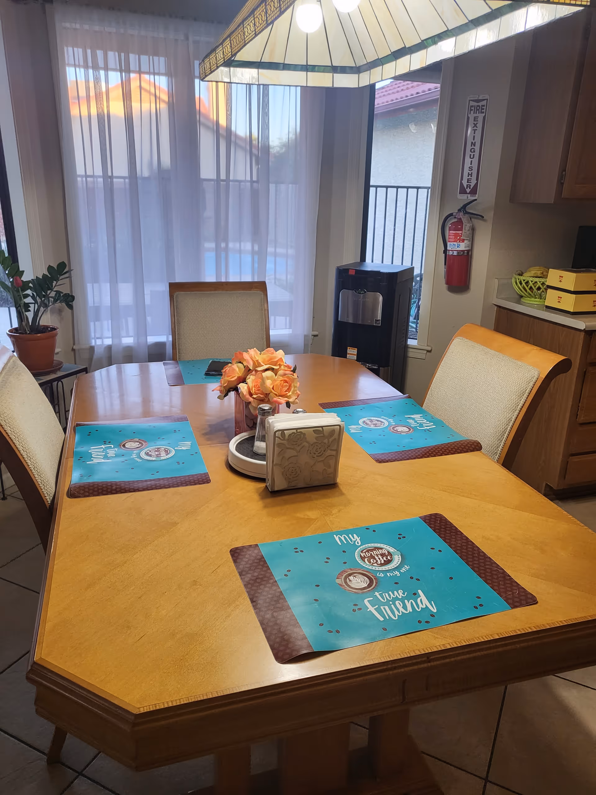 A dining area with a wooden table set for four with blue placemats that say 'My morning coffee is my best time friend.' There are four cushioned chairs around the table, a vase with orange flowers in the center, a napkin holder, and a salt and pepper shaker set. In the background, there is a window with sheer white curtains, a water dispenser, a fire extinguisher mounted on the wall, and kitchen cabinets with a basket of bananas on the counter.