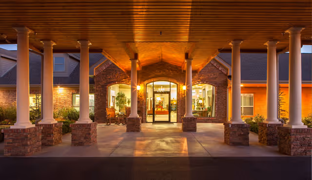 Entrance of a senior living facility at dusk with a covered driveway supported by white columns on brick bases, warm lighting illuminating the brick building and glass doors, and some plants and seating visible inside.