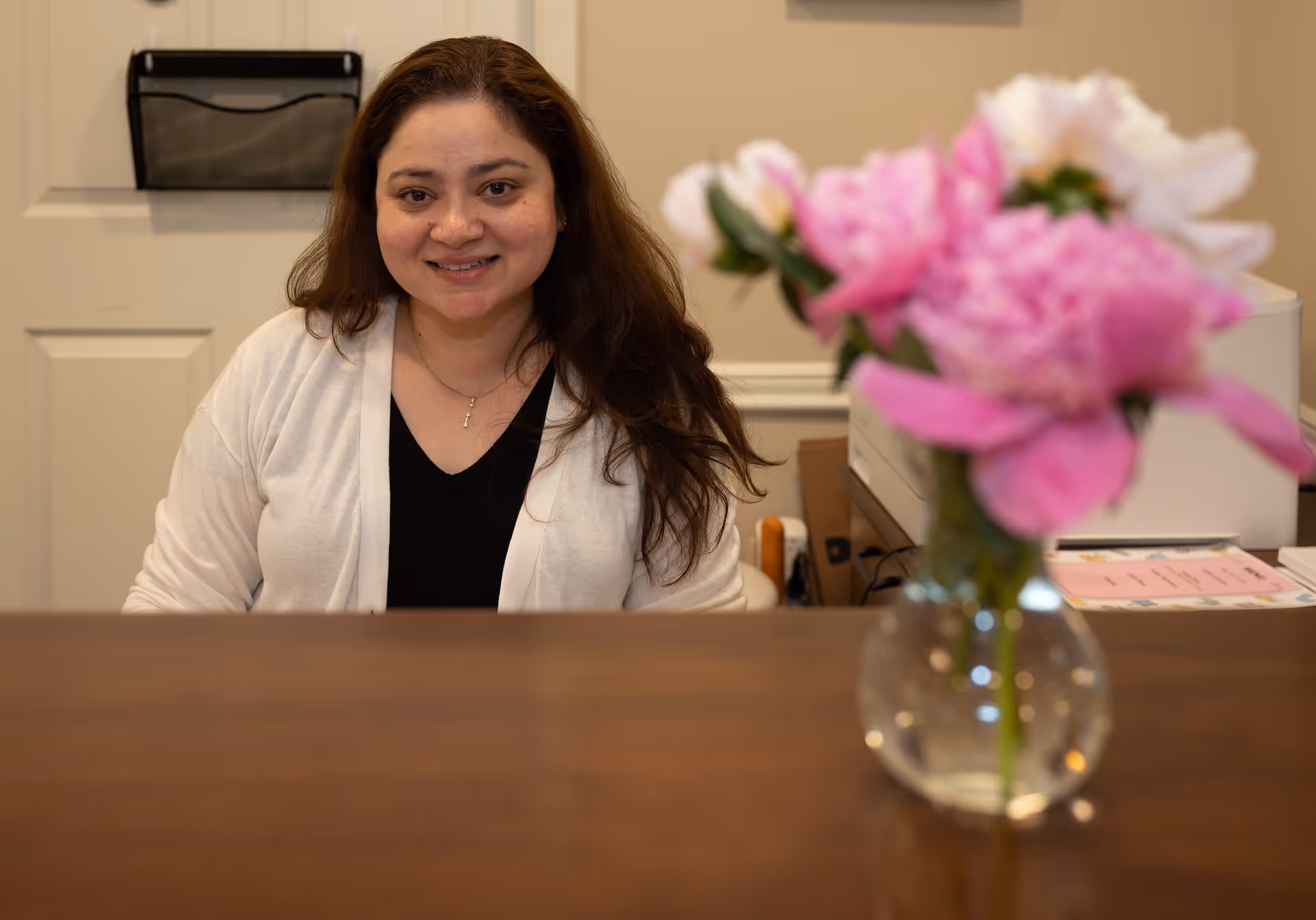 A woman with long brown hair wearing a white cardigan and black top is sitting behind a wooden desk. In the foreground, there is a glass vase with pink and white flowers. Behind her, there is a beige wall with a door and some office supplies.
