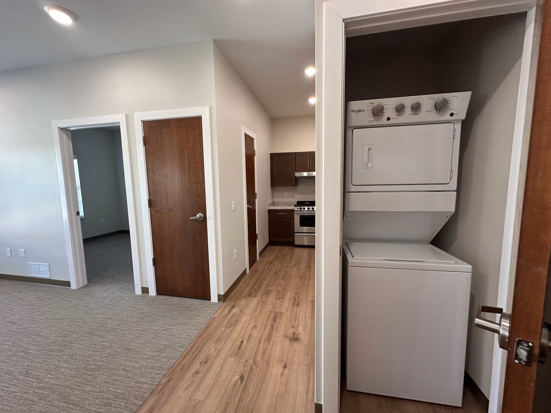 Interior view of a senior living apartment showing a laundry closet with a stacked washer and dryer on the right, a hallway with wooden flooring leading to a kitchen with dark cabinets and stainless steel appliances, and a carpeted room visible through an open doorway on the left.