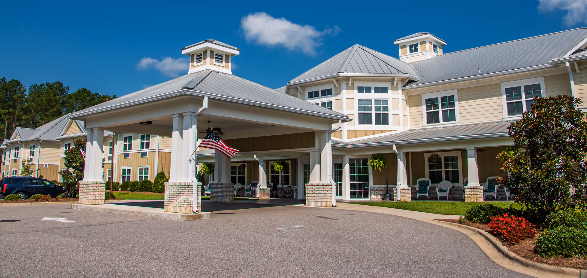 Exterior view of Arbor Landing at Ocean Isle, showing a large building with beige siding, white trim, and a metal roof under a blue sky with some clouds. The entrance has a covered drop-off area with white columns and brick bases, decorated with American flags. There are shrubs, flowers, and a few parked cars visible around the building.