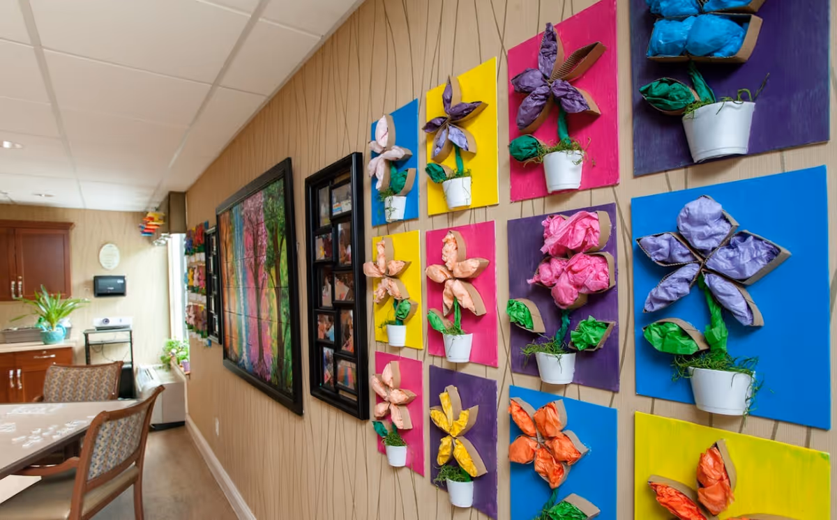 Interior view of a senior living facility hallway with colorful 3D flower art mounted on the wall. There is a dining table with chairs on the left side, wooden cabinets, and framed pictures on the wall. The space is well-lit with a ceiling light and has a warm, welcoming atmosphere.