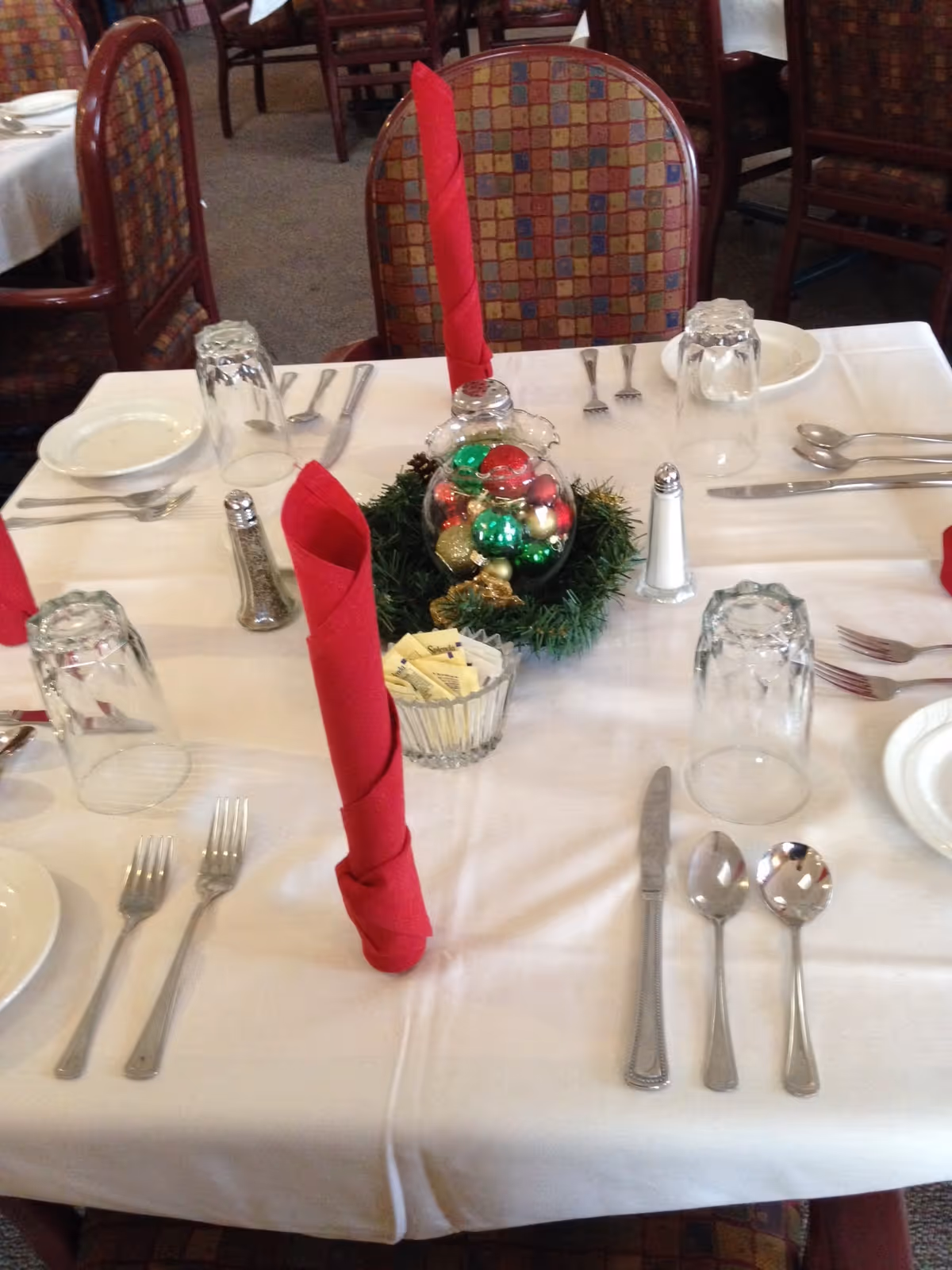 A dining table with a white tablecloth, folded red napkins, place settings and a festive glass ornament centerpiece.