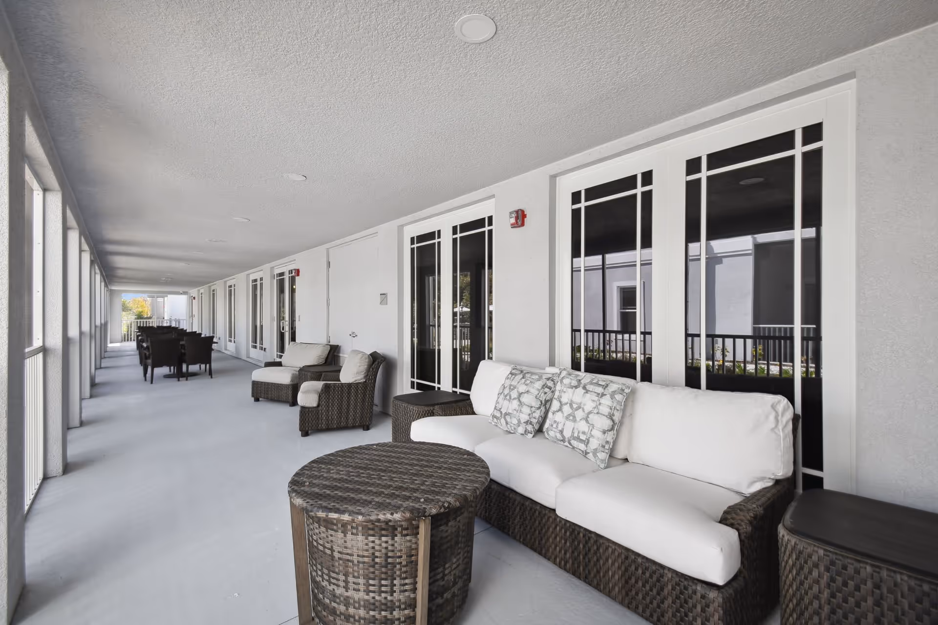 Covered outdoor corridor with wicker sofas, chairs and tables along a senior living facility balcony.