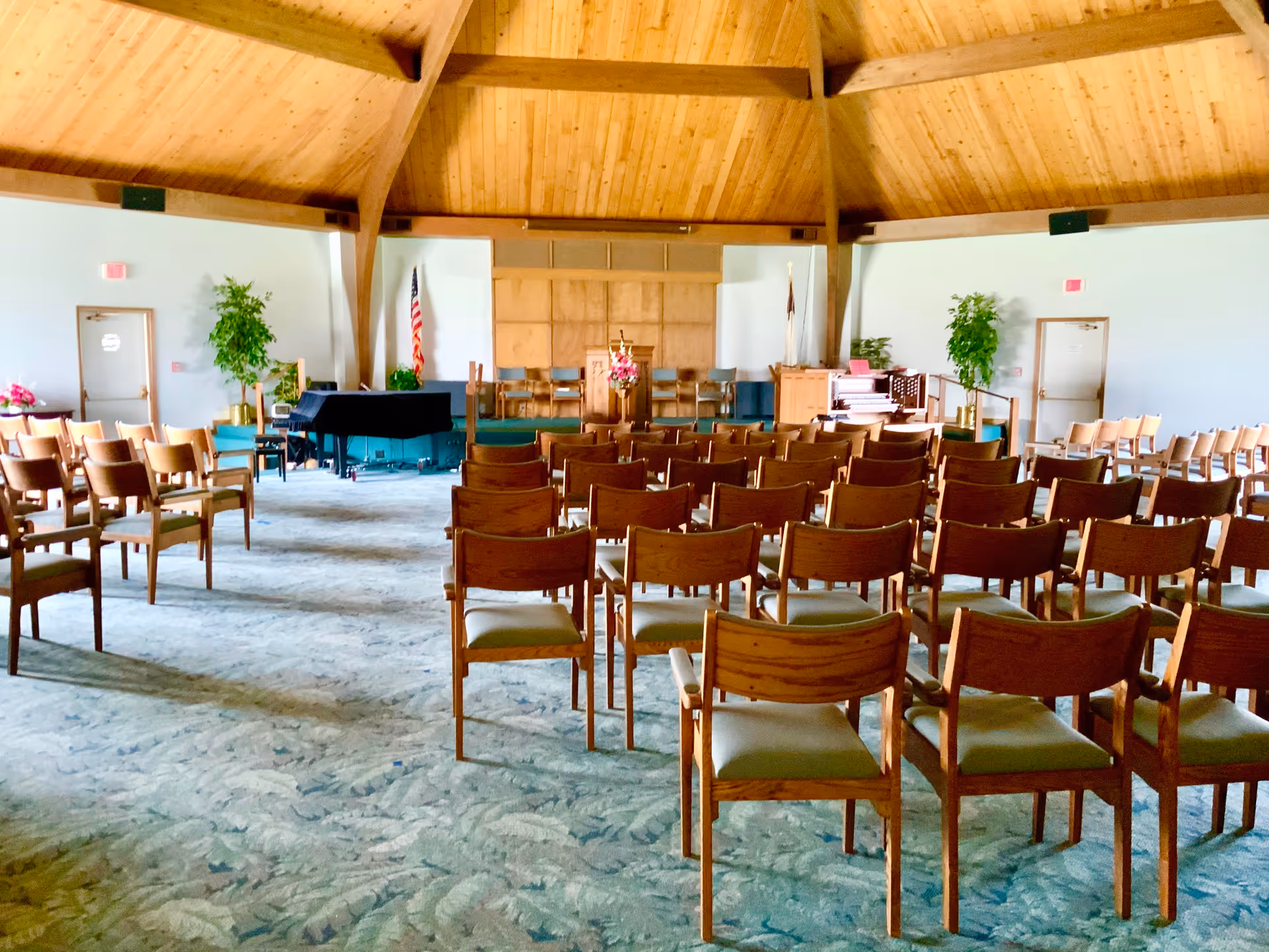 Interior view of a chapel or meeting room with rows of wooden chairs facing a stage area with a podium, piano, and organ. The ceiling is wooden with exposed beams, and there are two doors on either side of the room with potted plants nearby. American and Christian flags are displayed near the stage.
