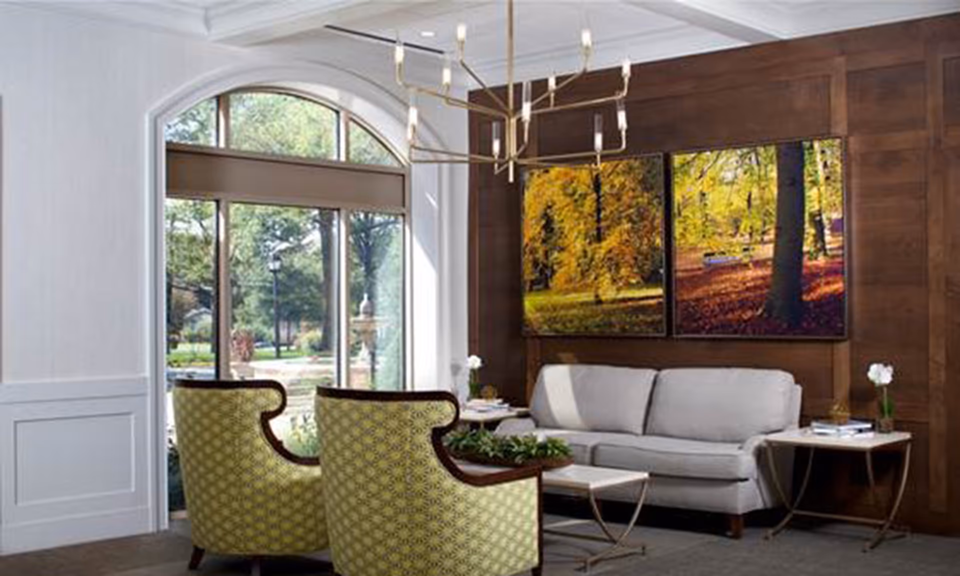 Bright seating area with a light gray sofa, two patterned green armchairs, a large arched window, wooden accent wall with landscape artwork, and a chandelier.