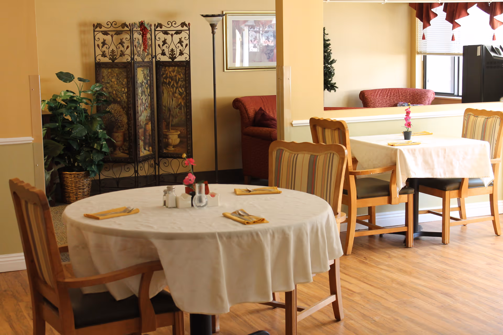 A dining area with two tables covered in white tablecloths, each set with yellow napkins and small flower centerpieces. Wooden chairs with striped cushions surround the tables. In the background, there is a decorative folding screen, a floor lamp, a potted plant, and a red upholstered sofa near a window with red valances.