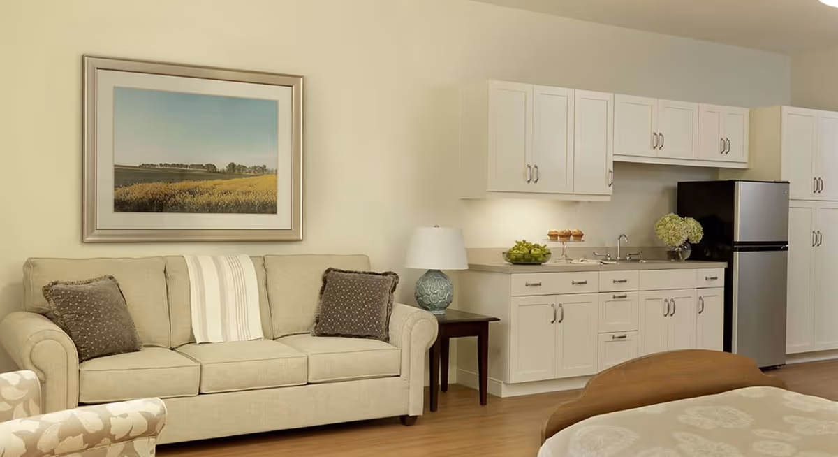 A cozy living area with a beige sofa adorned with two dark brown pillows and a white throw blanket. Next to the sofa is a small wooden side table with a decorative lamp. Behind the sofa, a framed landscape painting hangs on the wall. To the right, there is a kitchenette with white cabinets, a stainless steel refrigerator, a sink, and some decorative items including a bowl of green apples and a vase with flowers.