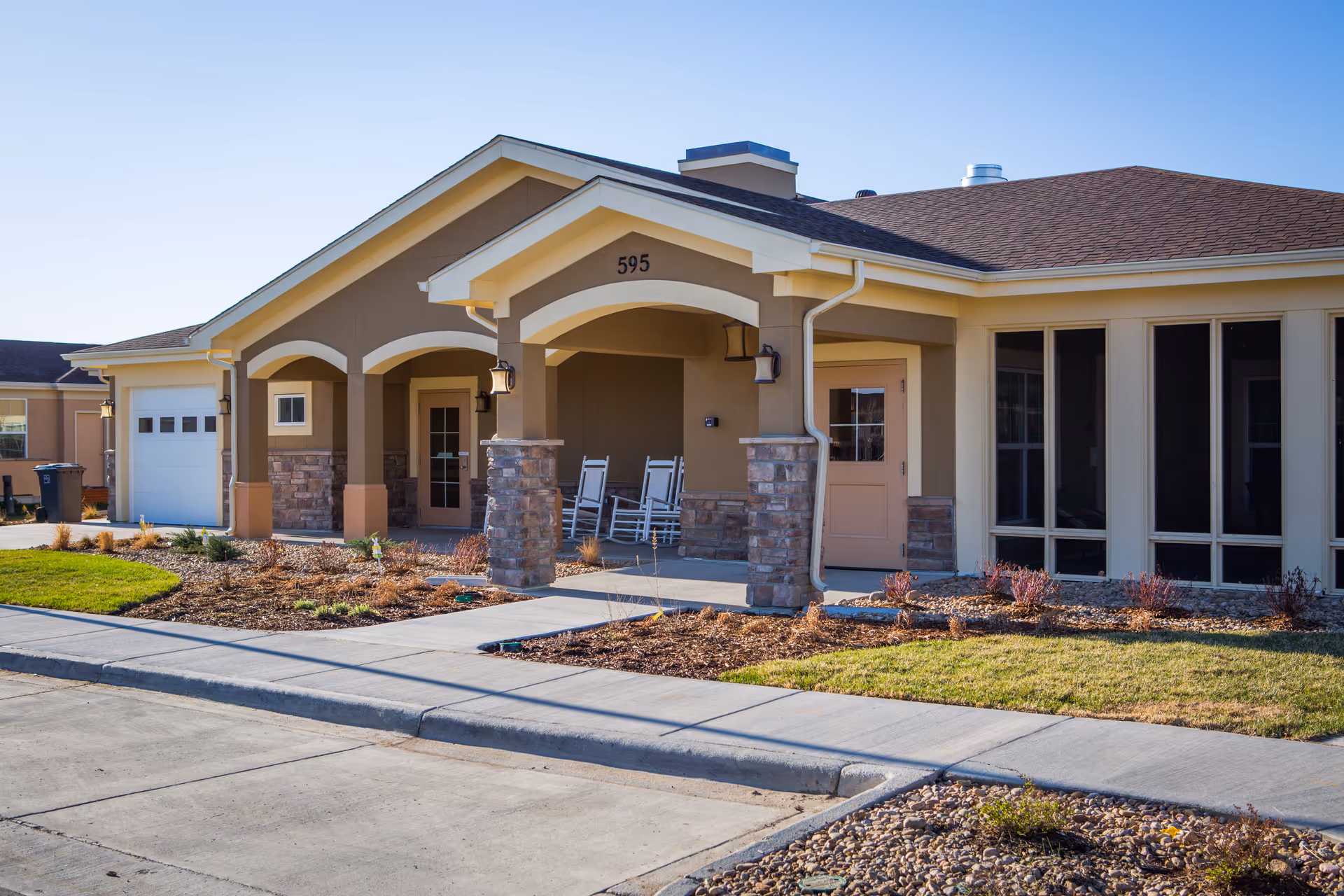 Front exterior of a single-story senior living home showing a covered entrance with stone pillars, rocking chairs, and the number 595.