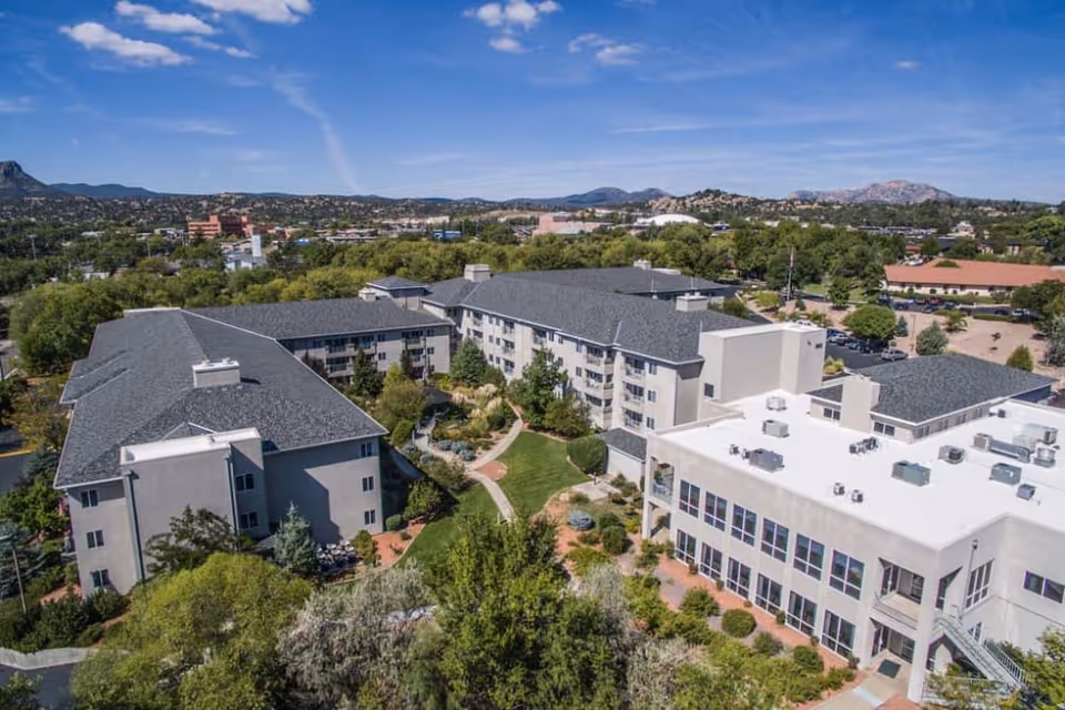 Aerial view of Las Fuentes Resort Village showing multiple connected buildings with gray roofs surrounded by trees and landscaped gardens, with mountains visible in the background under a blue sky.