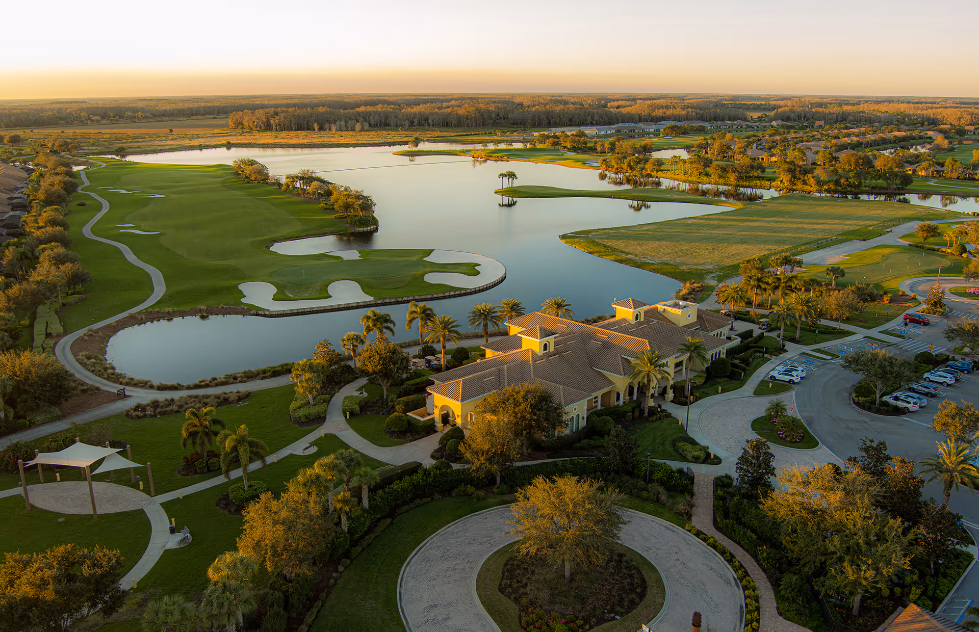 Aerial view of Del Webb Naples 55+ Retirement Community featuring a large clubhouse building surrounded by landscaped gardens, a circular driveway, parking lot with cars, a golf course with sand traps, water features, and walking paths under a clear sky during sunset.