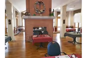 Interior view of a dining area with wooden floors, tables covered with red tablecloths, and chairs. A large brick fireplace with a decorative wreath is centrally located, dividing the room. The space is well-lit with natural light coming through windows.