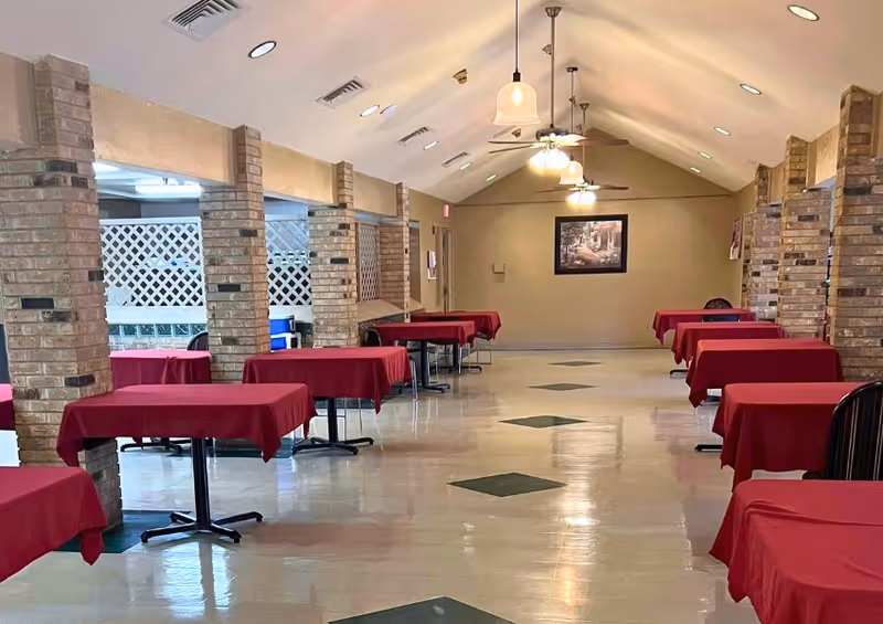 Long dining room with rows of small tables covered in red tablecloths, brick columns, and ceiling fans.