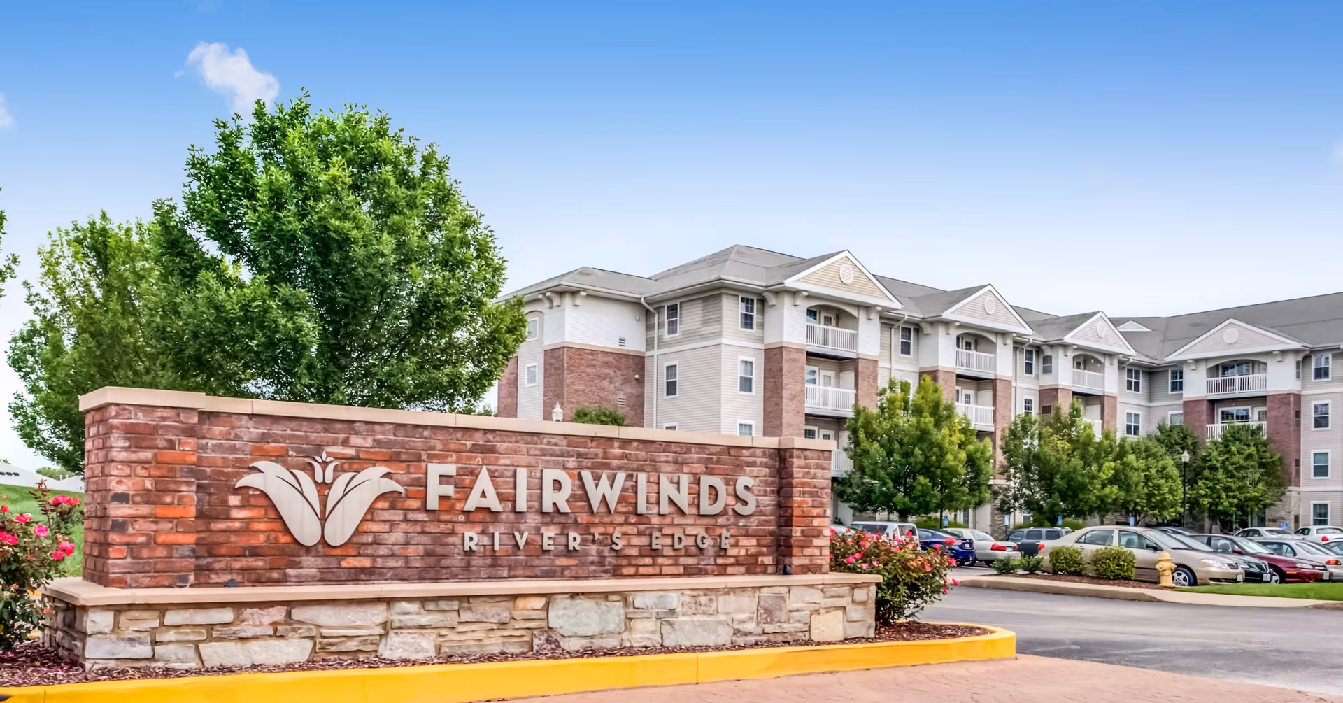 Exterior view of Fairwinds - River's Edge senior living facility showing a large brick sign with the facility name in front of a multi-story residential building surrounded by trees and parked cars under a blue sky.