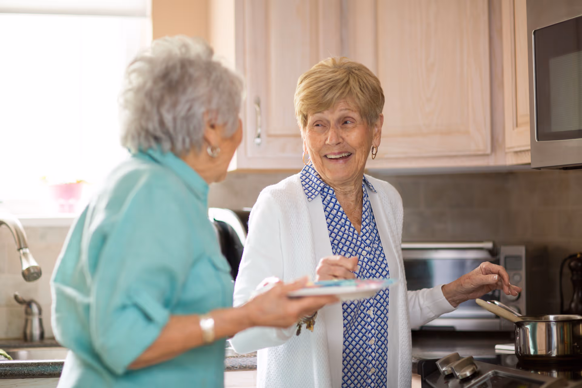 Two elderly women smiling and interacting in a kitchen. One woman is stirring a pot on the stove while the other holds a plate, and they appear to be enjoying each other's company.