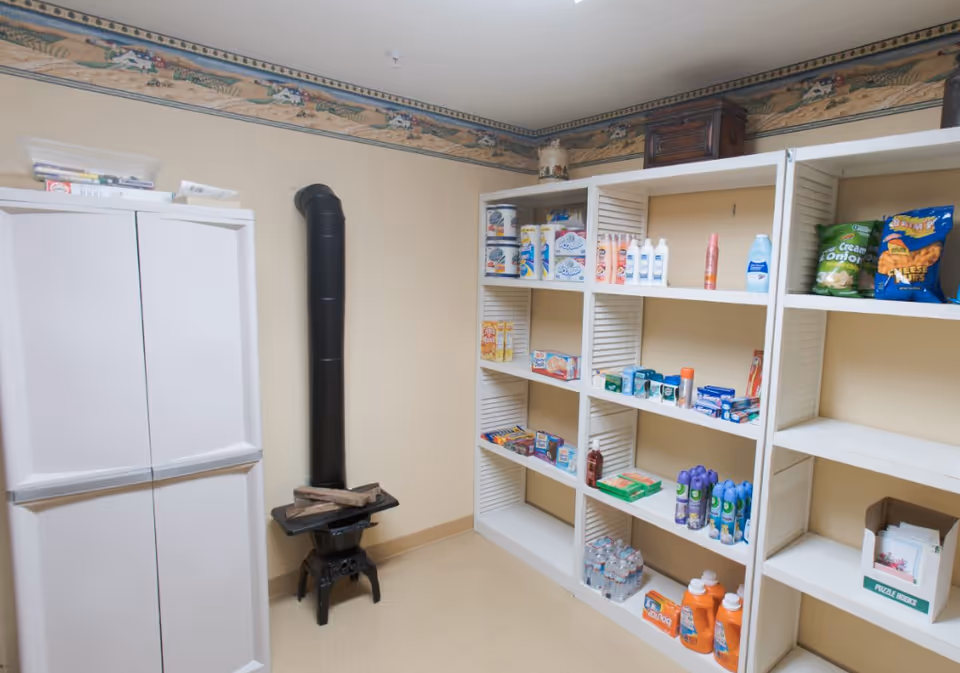 Small storage room with white shelving stocked with snacks, toiletries and cleaning supplies, a white cabinet, and a small black stove pipe.