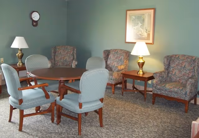 Cozy sitting room with a round wooden table surrounded by upholstered chairs, patterned armchairs, side tables with lamps, a wall clock, and framed artwork on green walls.