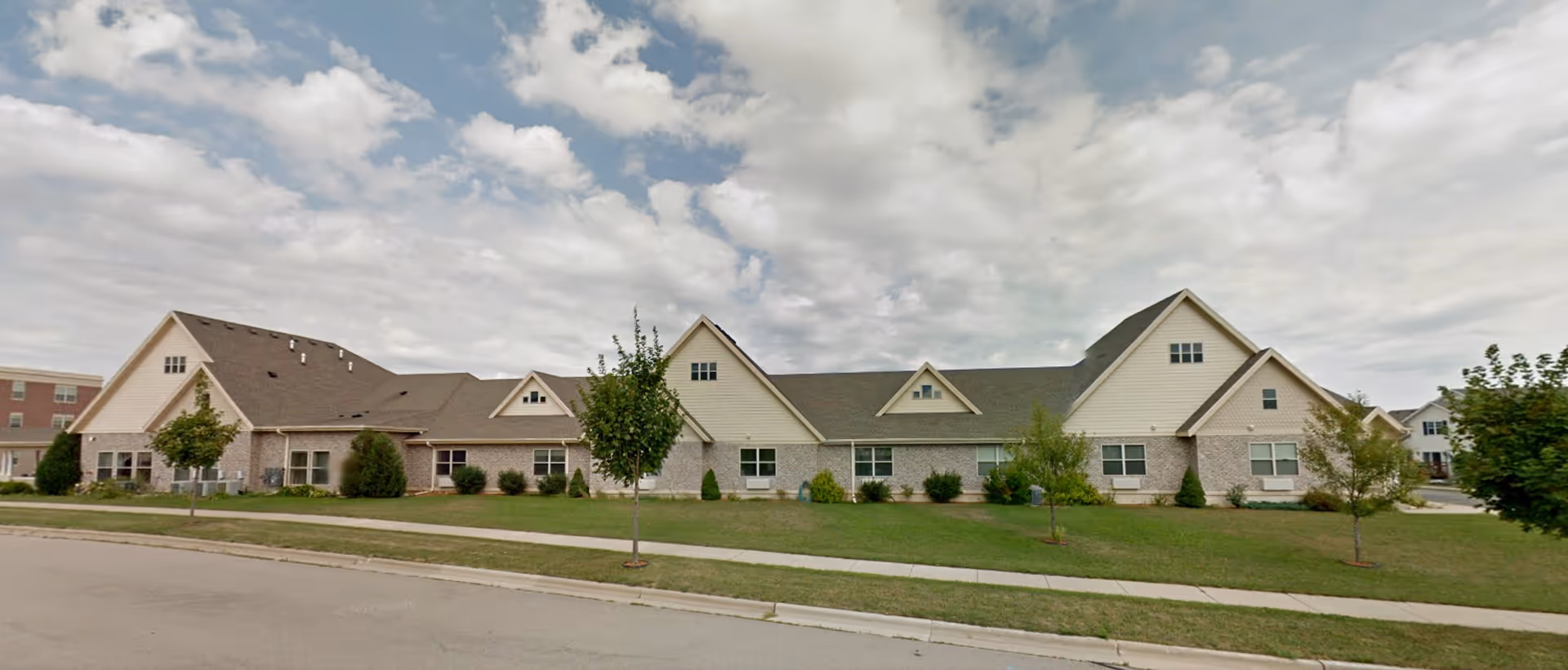 Exterior view of a single-story senior living facility building with a combination of brick and light-colored siding, multiple gabled roofs, several windows, small trees, and a grassy lawn under a partly cloudy sky.