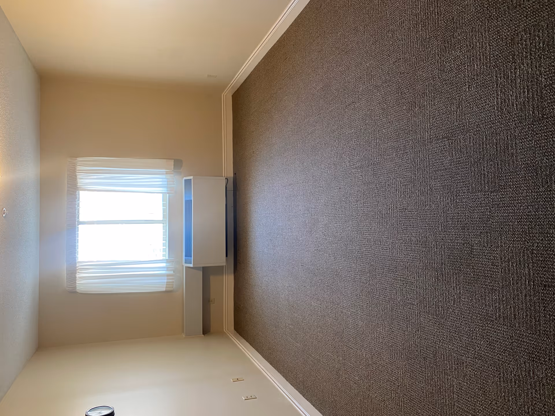 Empty carpeted living room with beige walls, a window with sheer curtains, and a small white cabinet beneath the window.