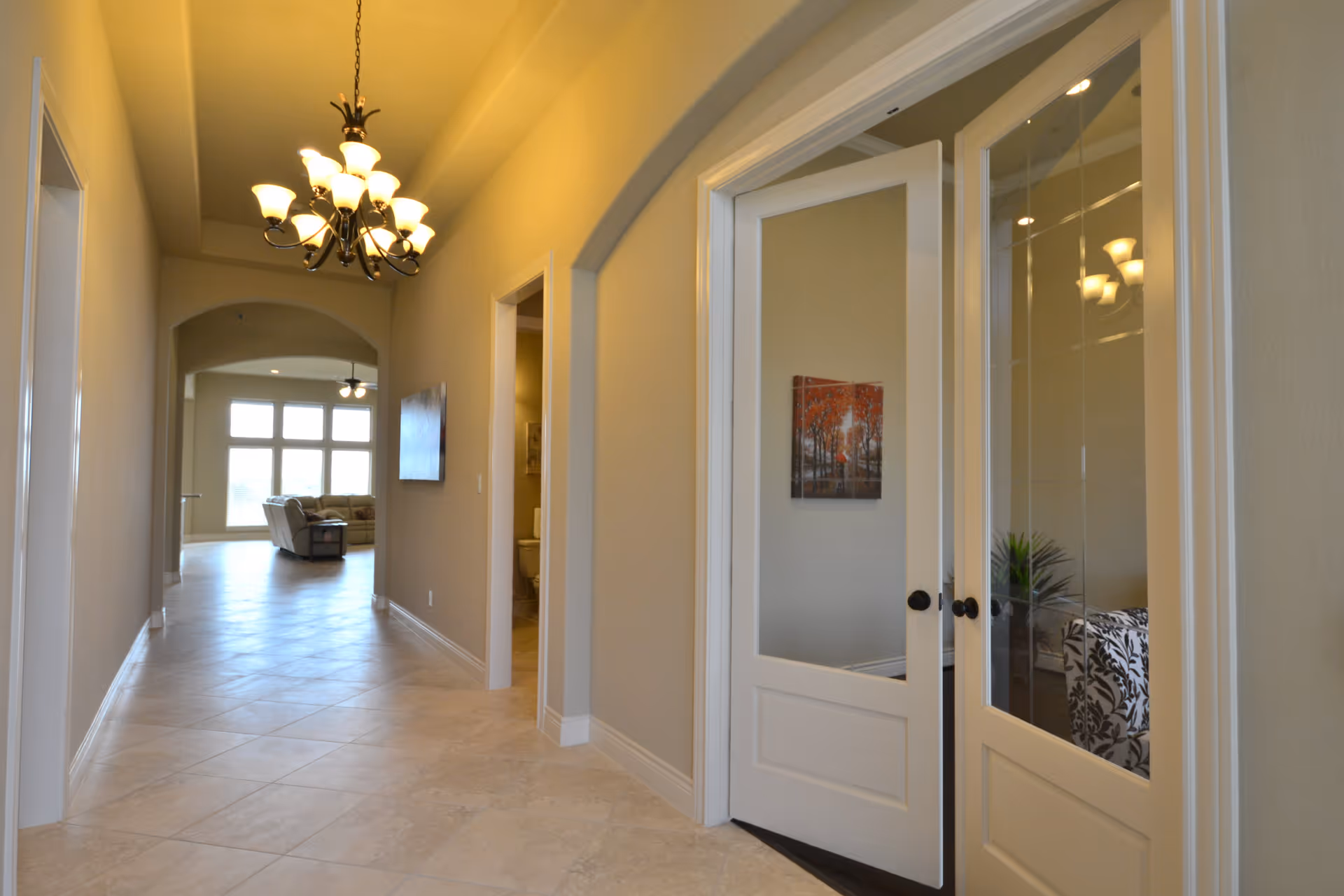 A well-lit hallway with beige walls and tiled floor leading to a living room area with large windows and a sofa. On the right side of the hallway, there are double glass-paneled doors with a painting and a plant visible inside the room. A chandelier hangs from the ceiling, and there is an open doorway to a bathroom on the right.