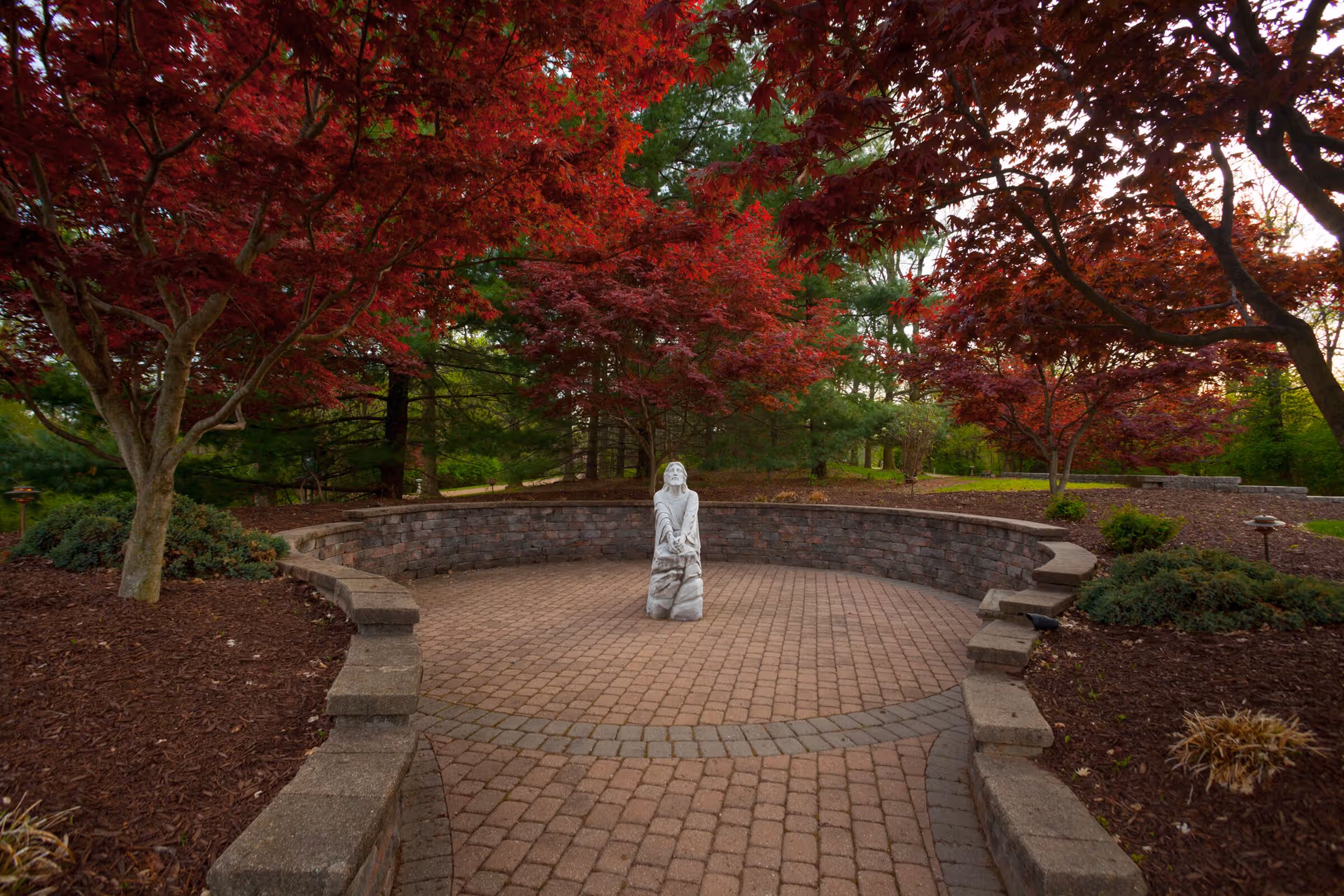A circular paved outdoor seating area with a low stone wall surrounded by red-leaved trees and green foliage. In the center of the paved area is a white statue of a person kneeling and looking upwards.