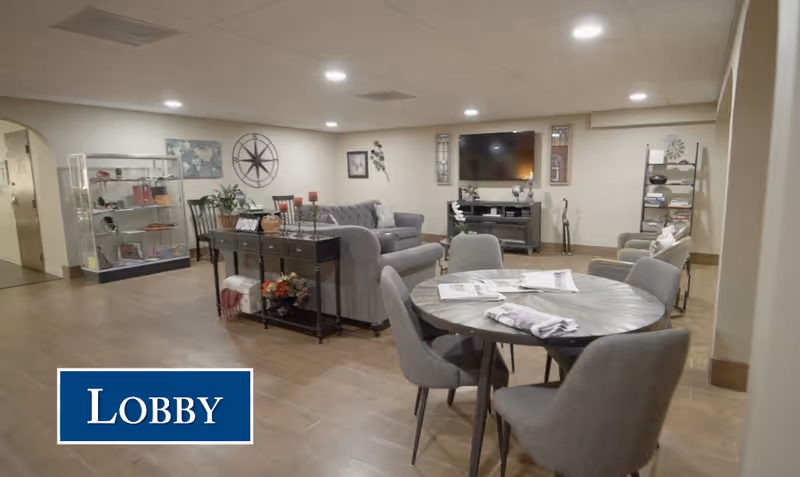 A spacious lobby area with a round table and four gray chairs in the foreground. Behind the table is a seating area with a gray sofa, two armchairs, and a TV mounted on the wall. The room is decorated with wall art, plants, and shelves with various items. The floor is wooden, and the ceiling has recessed lighting.