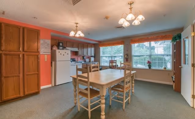 A dining area in a senior living facility with a wooden table and four chairs in the center. The room has large windows with orange valances letting in natural light. To the left, there is a kitchen area with wooden cabinets, a white refrigerator, and a stove. The walls are painted in a combination of coral and light blue colors. Two ceiling light fixtures with multiple bulbs illuminate the space.