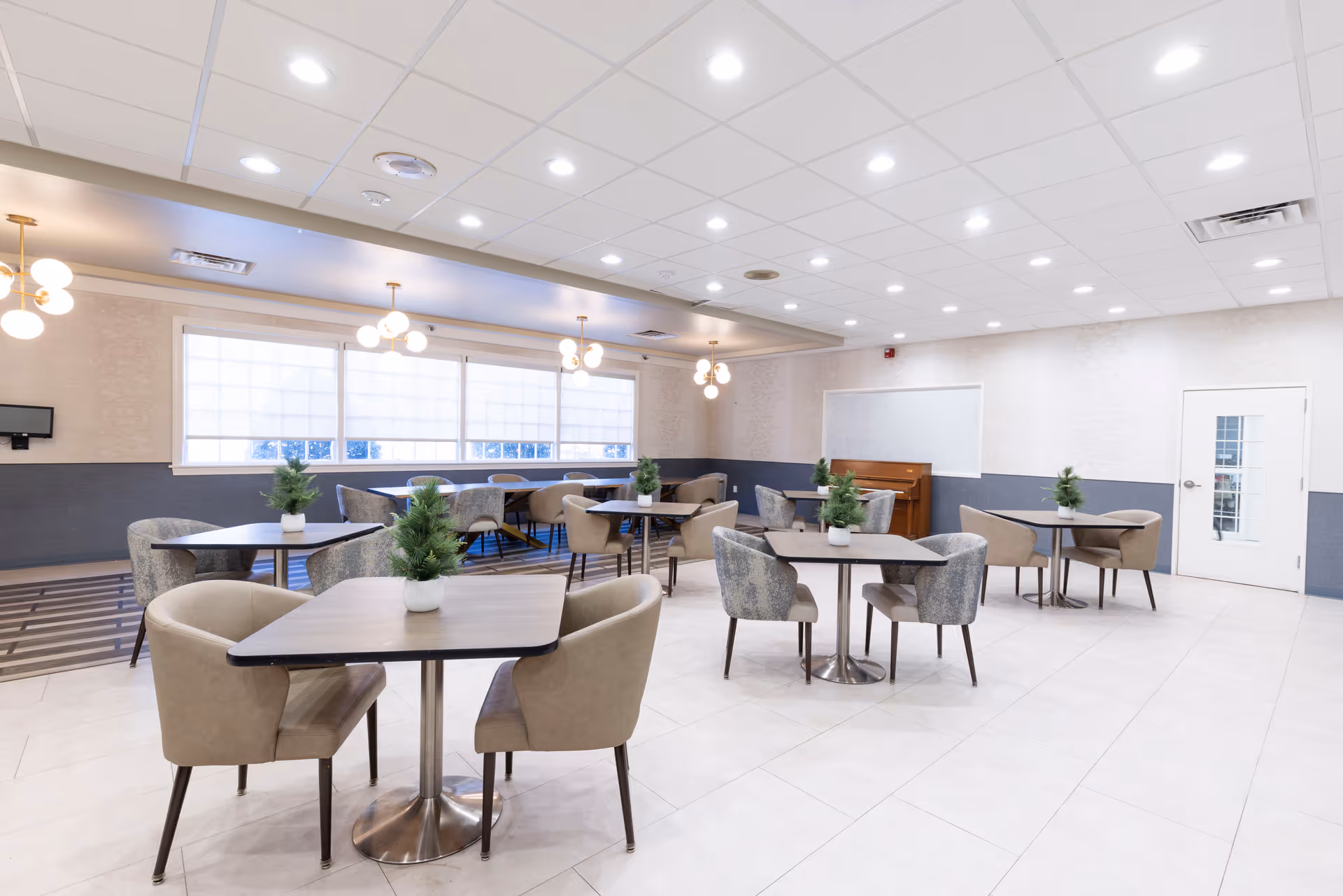 Bright communal dining room with several small tables and upholstered chairs, potted plants on each table, and a piano along the back wall.