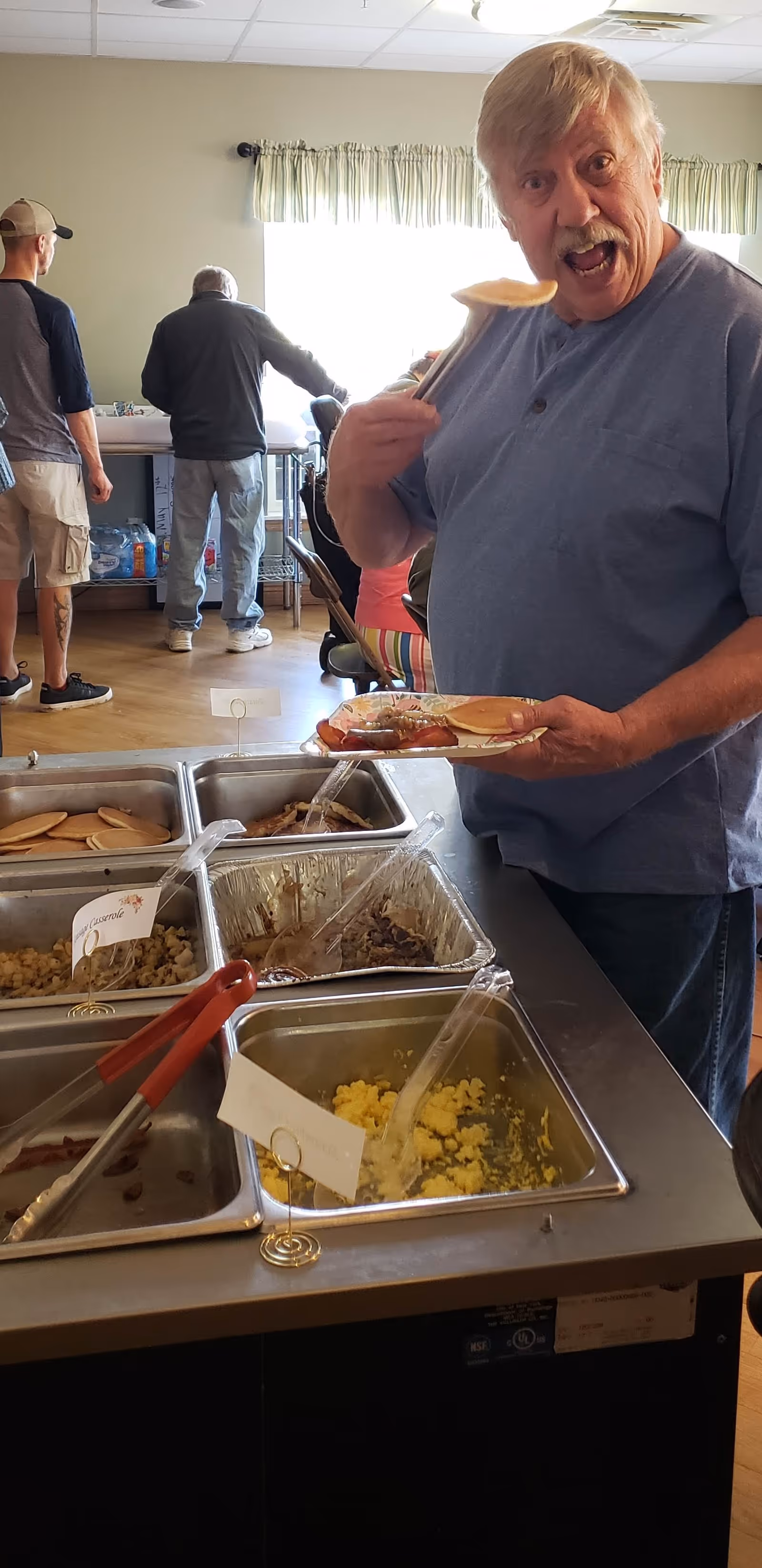 An elderly man in a blue shirt is serving himself food from a buffet-style setup with trays containing scrambled eggs, pancakes, and other breakfast items. He is holding a plate and a serving utensil, smiling with an open mouth. In the background, two other men are standing near a counter with a window covered by light green curtains.
